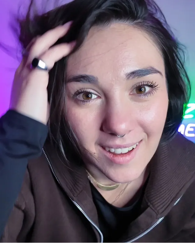 Mel in primo piano mentre si sistema i capelli, con luci al neon viola e verdi alle spalle.